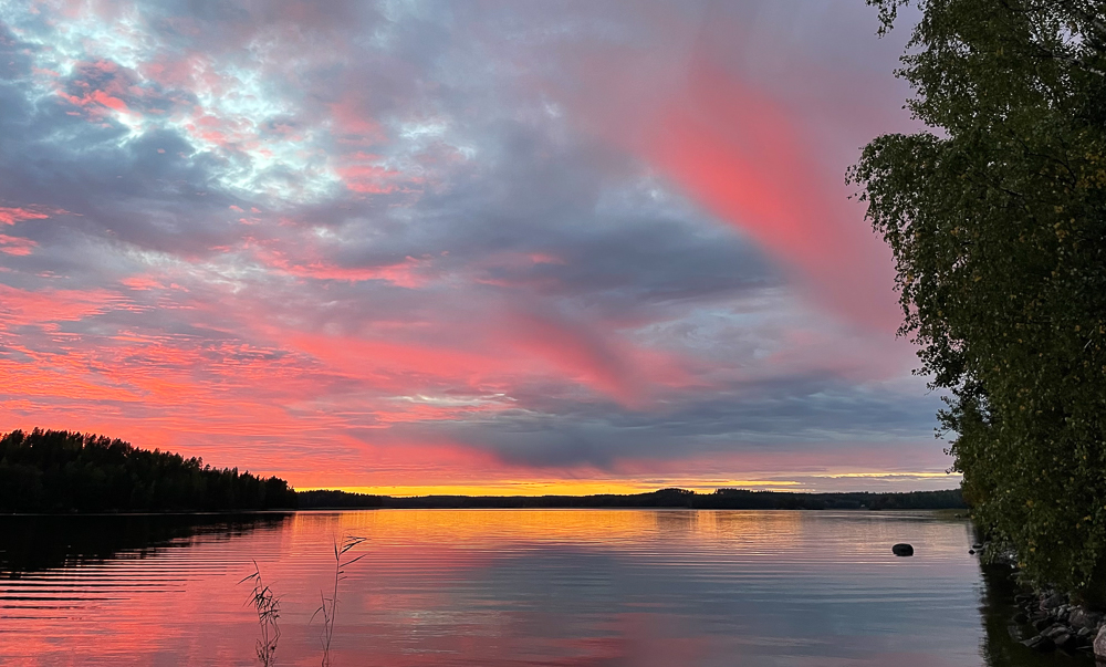 Punakeltainen auringonlasku järven takana ja punaisia pilviä taivaalla.