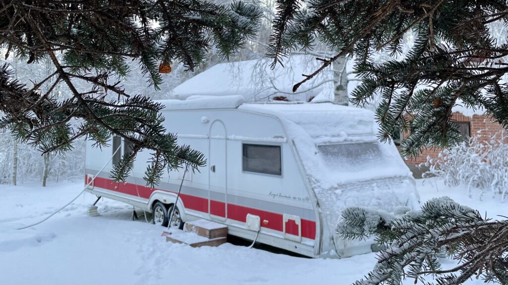 Etualalla olevien kuusen oksien takaa näkyy Kabe-asuntovaunu lumisella pihamaalla.